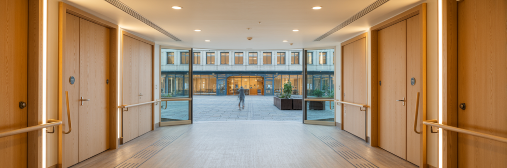 Couloir courbe d’un EHPAD moderne avec portes en chêne clair, éclairage LED, plantes et salon commun au fond.